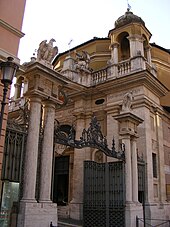 Farbfotografie in Untersicht vom Eisentor und Eingangsportal einer Kirche, die von Adlern auf Säulen begrenzt ist. Die Kirche besteht aus einem Säuleneingang, einem Eckturm und einem runden Mittelteil.