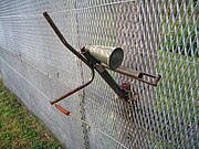 Horn-shaped device mounted on the side of a metal fence, with trigger wires attached to it and running parallel to the fence into the foreground and background.