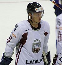 An ice hockey player standing, looking to the right of the camera. He is wearing a black helmet and a white uniform.