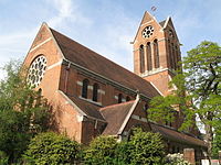 A late Victorian brick church with a gabled tower seen above a collection of trees.