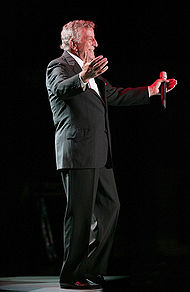 Tony Bennett is opening his arms, and holding a mic in his hand.