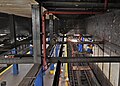 Blick von oben in einen U-Bahnhof. Das dunkel gestrichene Stahlskelett mit Stützen und Trägern ist gut sichtbar. In der rechten Bildhälfte fliegen Funken, weil zwei Arbeiter auf einem fahrbaren Baugerüst stehend den Anstrich abschleifen.