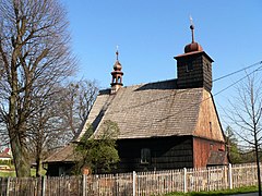 Zdjęcie wykonane w słoneczny dzień przedstawia drewniany kościół w Rzepiszczu, w ujęciu z boku. Fotografia wykonana jest z drogi, na pierwszym planie jest drewniane ogrodzenie. Kościół jest niewielki, dominującym jego elementem jest wysoki dach pokryty gontem. Z dachu wyrastają dwie wieże - większa nad wejściem i mała, tzw. sygnaturka, nad prezbiterium. Obok kościoła rośnie drzewo, na zdjęciu jest bez liści.