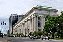 A tall white stone building with a colonnaded facade and intricate decorations on the stonework, much longer along the street to its left then the side facing the camera. There are trees in front of it on the right and a taller, more modern building behind it.