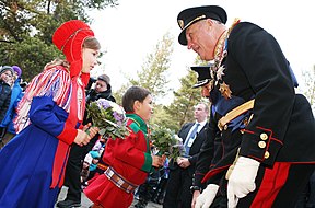 Photograph of the king of Norway speaking to and receiving a bouquet of flowers from a young girl dressed in a traditional Sami gaeptie