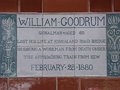 A tablet formed of six standard sized tiles, bordered by green flowers in the style of the Arts and Crafts movement. The tablet reads "William Goodrum signalman aged 60 lost his life at Kingsland road bridge in saving a workman from death under the approaching train from Kew February 28, 1880".