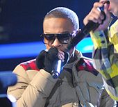 A picture of a man wearing glasses while holding a microphone in their hands wearing a thick jacket and gloves