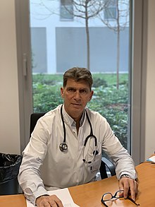 Photograph of Philippe Halfon in a white coat, sat at a desk in an office