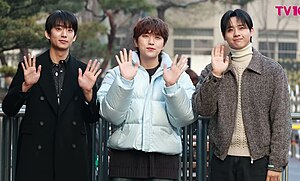 Gongchan, Sandeul, and CNU of B1A4 standing at Music Bank, waving and smiling at the camera. Gongchan is wearing a long black coat, Sandeul is wearing a pale blue puffer coat, and CNU is wearing a brown knit zip up jacket.