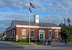 A low brick building with a square cupola in the middle of its metal roof. Wording on the front says "UNITED STATES POST OFFICE" and "ALBION NEW YORK". There is an American flag flying from a flagpole in front and hanging from a lamppost on the right.