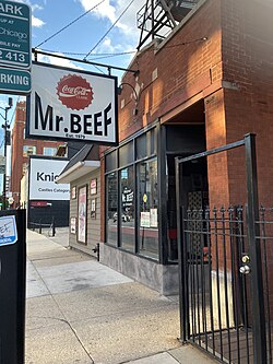 A large white sign with the words "Mr. Beef" and a Coca-Cola logo hangs over a sidewalk. The building of the restaurant is brick with a glass window in the front.