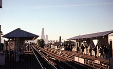 A train station with two wooden side platforms, each with gooseneck lights and hipped roof canopies, is seen looking southeast. Partially clouded by fog, the Sears Tower is also visible.