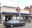 A white-bricked building with a rectangular, dark blue sign reading "WEST KENSINGTON STATION" in white letters all under a light blue sky