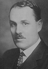 Black and white photo of a middle-aged man with a small moustache, wearing a dark suitcoat, white dress shirt, and a white-spotted necktie