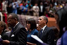 Obama and his wife standing in a crowded Church, looking forward, with their mouths open mid-sentence while reciting a prayer.