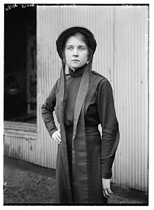 A young white woman, standing outdoors, wearing dark plain dress and bonnet