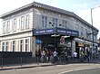 A white-bricked building with a rectangular, dark blue sign reading "WILLESDEN GREEN STATION" in white letters all under a light blue sky