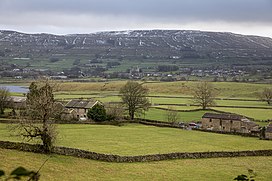 Green fields in the foreground with a mountain range in the background scattered with snow