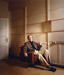 Richard Ford sitzt auf einer Couch und hält die Faust hoch, hinter ihm eine eingeschlagene Wand.