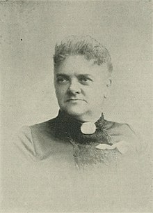 B&W portrait photo of a woman with her hair in an updo, wearing a dark blouse with a white ribbon pinned on it, as well as a white brooch