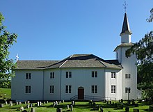 Foto einer weißen Holzkirche. Rechts im Bild der Kirchturm, daran schließt sich ein achteckiger Gebäudeteil an, danach ein Gebäudeteil mit rechteckigem Grundriss. Vor der Kirche befinden sich Grabsteine