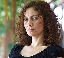 Head shot of Maha Haj, she has shoulder-length curly brown hair. The background is blurry trees