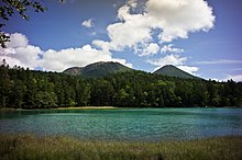 Am Gras bewachsenen Ufer stehender Blick über den kleinen See. Auf der gegenüberliegenden Seite befindet sich dichter Wald aus dem im Hintergrund zwei niedrigere, bewachsene Berge herausragen.