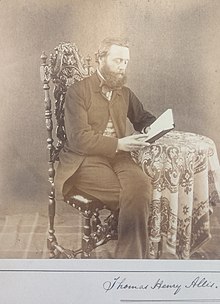 Bearded man in a suit, posed sitting at a table looking at a book