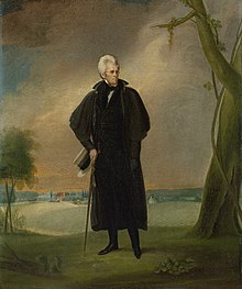 White-haired man stands outdoors beside a tree with cane.