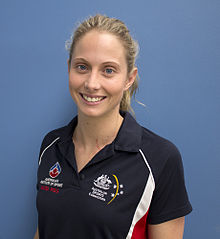 Glencora Ralph trägt ein dunkelblaues Hemd und steht vor hinreichend kontrastiertem, hellerem blauen Hintergrund. Auf dem Hemd befinden sich die Logos des Australian Institute of Sport und der Australian Sports Commission.