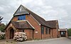 Three-quarter view of a modern brick building in a vernacular style, resembling a cottage. A porch with an arched entrance projects from the façade; above its brown-tiled roof is a three-light window and some wooden panelling with a white crucifix. Three evenly spaced side windows and a larger round-arched window extend down the side.