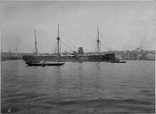 A large gray ship with three masts and a wide smoke stack at anchor; several small boats lie in front.