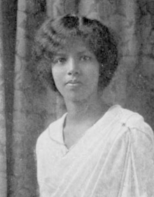 A young, dark-skinned South Asian woman, wearing a white dress. Her dark hair is parted and dressed back and low at the nape. She is looking directly at the camera and not smiling.