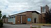 A low, modern brick building with an entrance door surrounded by a ground-to-roof glazed wall. A square tower with a metal-clad glazed area and a crucifix stands to the right. A low-roofed extension to the left has tall windows on one side and one narrow window on the other.