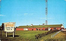 A one-story brick building set far back from the street, with a large transmitter tower visible behind it. In front is a sign reading "Television Manor, K H O L - T V, Channel 13".