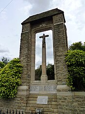 A photograph of a tall stone monument with a crucifix in the centre and a small V-shaped roof supported by two pillars. The names of those who fell in the two world wars are carved in stone at the base of the monument