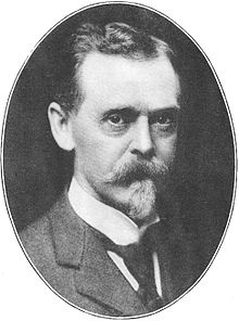 Black-and-white photograph, set in an oval frame, of the head and shoulders of a man of about 55. Dressed formally in a dark suit, dark tie, and white collar, he is looking directly out of the frame. He has dark eyes and dark hair, but his neatly trimmed moustache and goatee are gray.