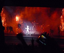 Faraway image of a stage lit by potlight falls on a female blond woman in the centre who wears a large white headdress and holds a stick in her left hand. She is surrounded by similar dressed male dancers who also wear a half-sleeved jacket.