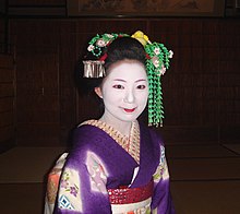 A maiko wearing a purple kimono and a long green hair ornament on her left side.