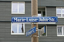 Blaues Straßenschild mit weißer Schrift "Marie-Luise-Jahn-Str.". Das Schild ist an einem runden Holzpfahl angebracht, dahinter sieht man ein graues Gebäude.