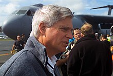 Image of Ambassador Swindells, right side profile, from the shoulders up. He's wearing a grey fleece jacket. Behind him, out of focus, are air force personnel and a grey C-141 Starlifter cargo plane.