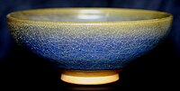 A ceramic bowl is viewed from one side. The glaze has a crazed and mottled finish, varying from yellow at the rim of the bowl to blue below.