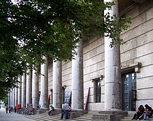 Die massive Fassade in diagonaler Ansicht, mit der Reihe der Säulen, der Freitreppe und einigen Ästen der Linden vor dem Gebäude