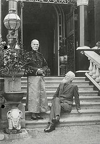 outdoor shot with two men on steps of a large house: bearded Western men (seated, right) wearing suit; one oriental-looking man (left, standing) wearing Chinese garments