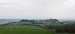 Farmland sloping gently away and then rising towards a ridge. The remains of the hill fort, which sits on the ridge, is obscured by trees.