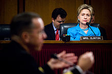 Clinton listens as the Chief of Naval Operations, Admiral Michael Mullen, responds to a question during his 2007 confirmation hearing with the Senate Armed Services Committee. She is in the background, sitting behind a desk with a placard bearing the words "MRS CLINTON", and is wearing a blue suit. A man wearing a black suit sits behind Clinton, taking notes.