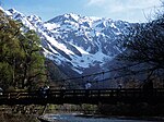 A suspension bridge over a river with high snowcovered mountains in the back.