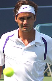 A brown-haired man in a white polo shirt moves towards the camera, as a ball can be seen bouncing in the foreground