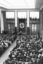 Die Mittelhalle voll mit Besuchern in Sitzreihen, an der Schmalseite spricht Adolf Hitler an einem Rednerpult vor einer gigantischen Hakenkreuzfahne