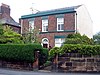 A sandstone wall with a hedge and a gate in front of a plain brick house in two storeys with three bays and an arched central doorway; the right side of the house is painted white and the edge of a larger house is to the left